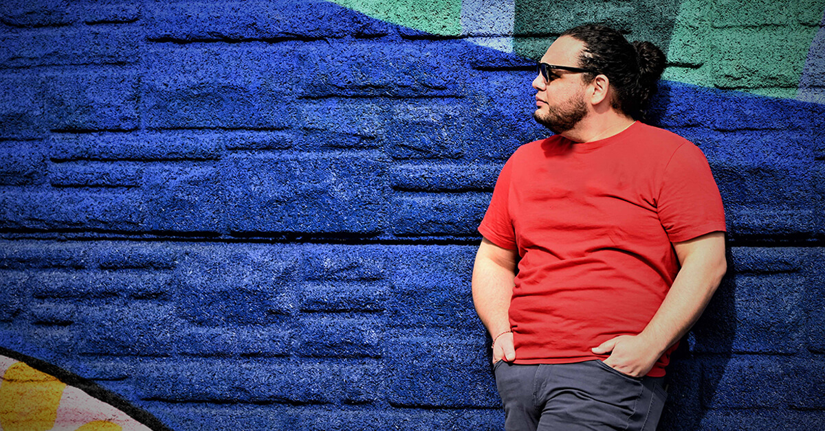 Capital One associate Aaron stands against a colorful wall and looks off, wearing a red shirt